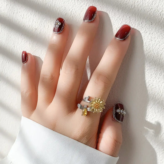 Bague florale ornée de détails rouges et dorés portée sur une main avec du vernis à ongles bordeaux pailleté.